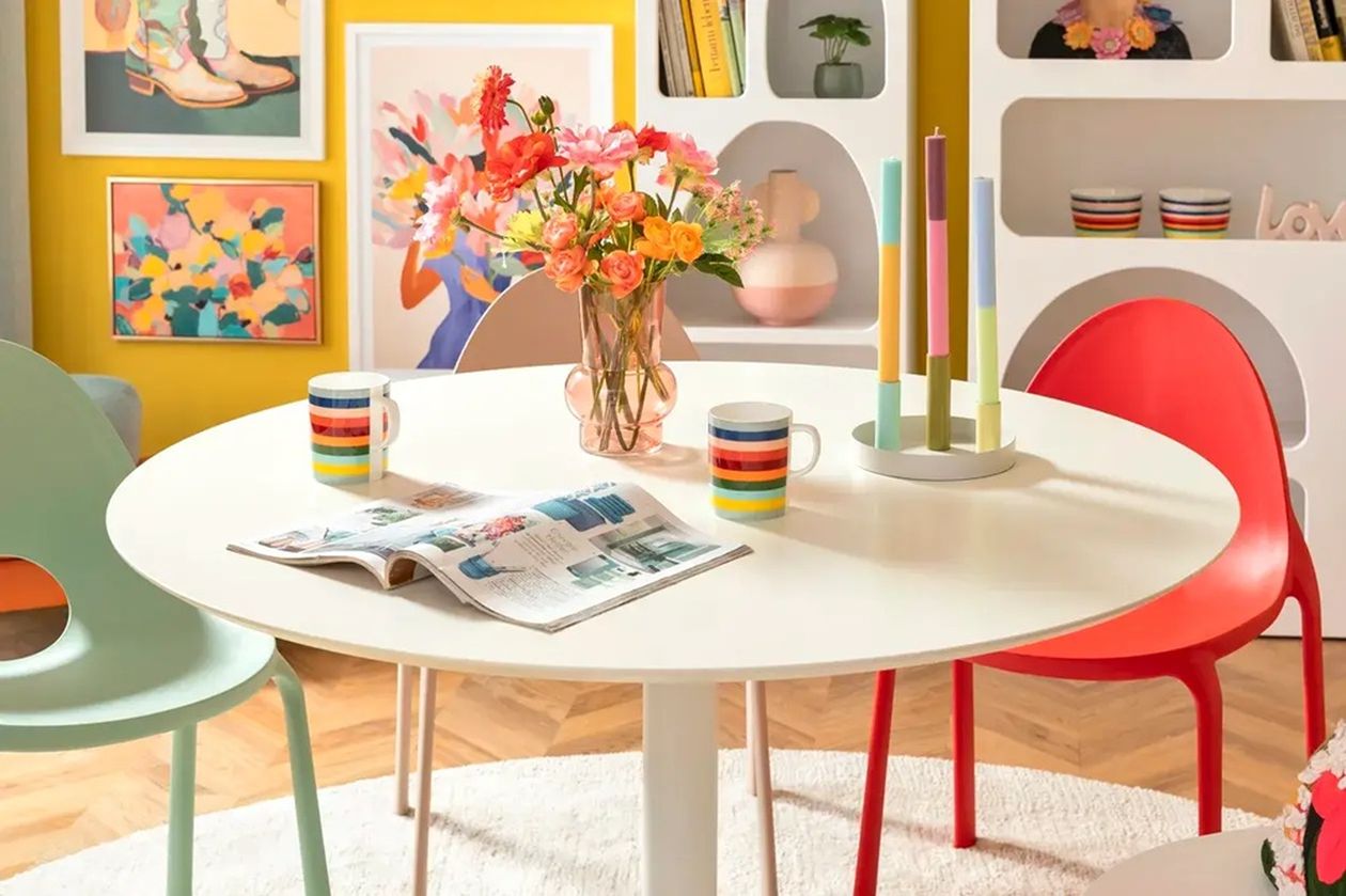 Runder, weißer Esstisch mit bunten Stühlen in einem lebendig gestalteten Raum. Auf dem Tisch stehen ein Strauß frischer Blumen, bunte Kerzen und Tassen mit Streifen. Im Hintergrund bunte Wandkunst und offene Regale.