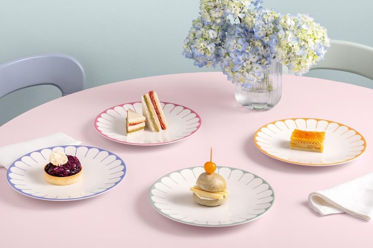 Vier weiße Frühstücksteller mit farbigem Muschelrand (pink, blau, grün, orange) auf rosa Tisch, jeweils mit kleinen Snacks belegt; im Hintergrund eine Glasvase mit blauen Hortensien.