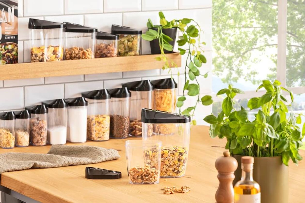 Moderne Küche mit zahlreichen transparenten Vorratsdosen mit schwarzen Deckeln, randvoll mit Lebensmitteln. Dazwischen stehen grüne Kräuter und verschiedene Gewürze.