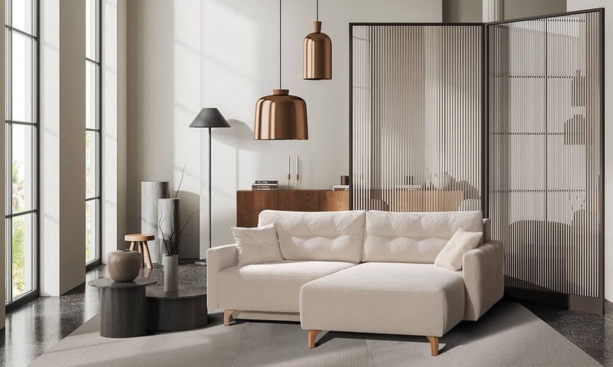 Minimalistisches Wohnzimmer mit cremefarbenem Ecksofa mit Récamiere rechts und hellen Holzfüßen. Im Hintergrund ein Paravent als Raumteiler, kupferfarbene Pendelleuchten und eine schwarze Stehleuchte. Der Raum hat große Fenster und einen dunklen Terrazzoboden.