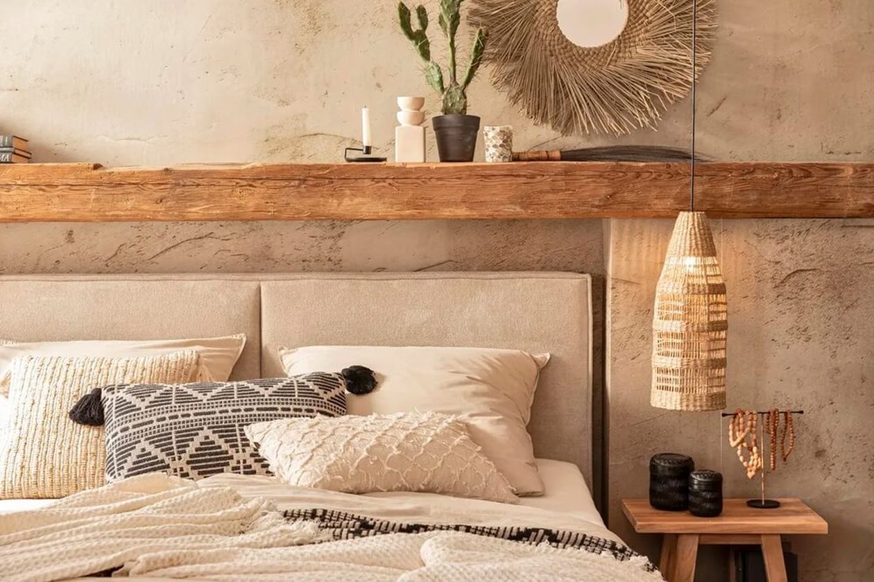 Beigefarbenes Polsterbett mit gepolstertem Kopfteil in einem Schlafzimmer im Boho-Stil. Über dem Bett ein Holzbalken als Ablage mit Deko und eine Flechtlampe als Hängeleuchte.