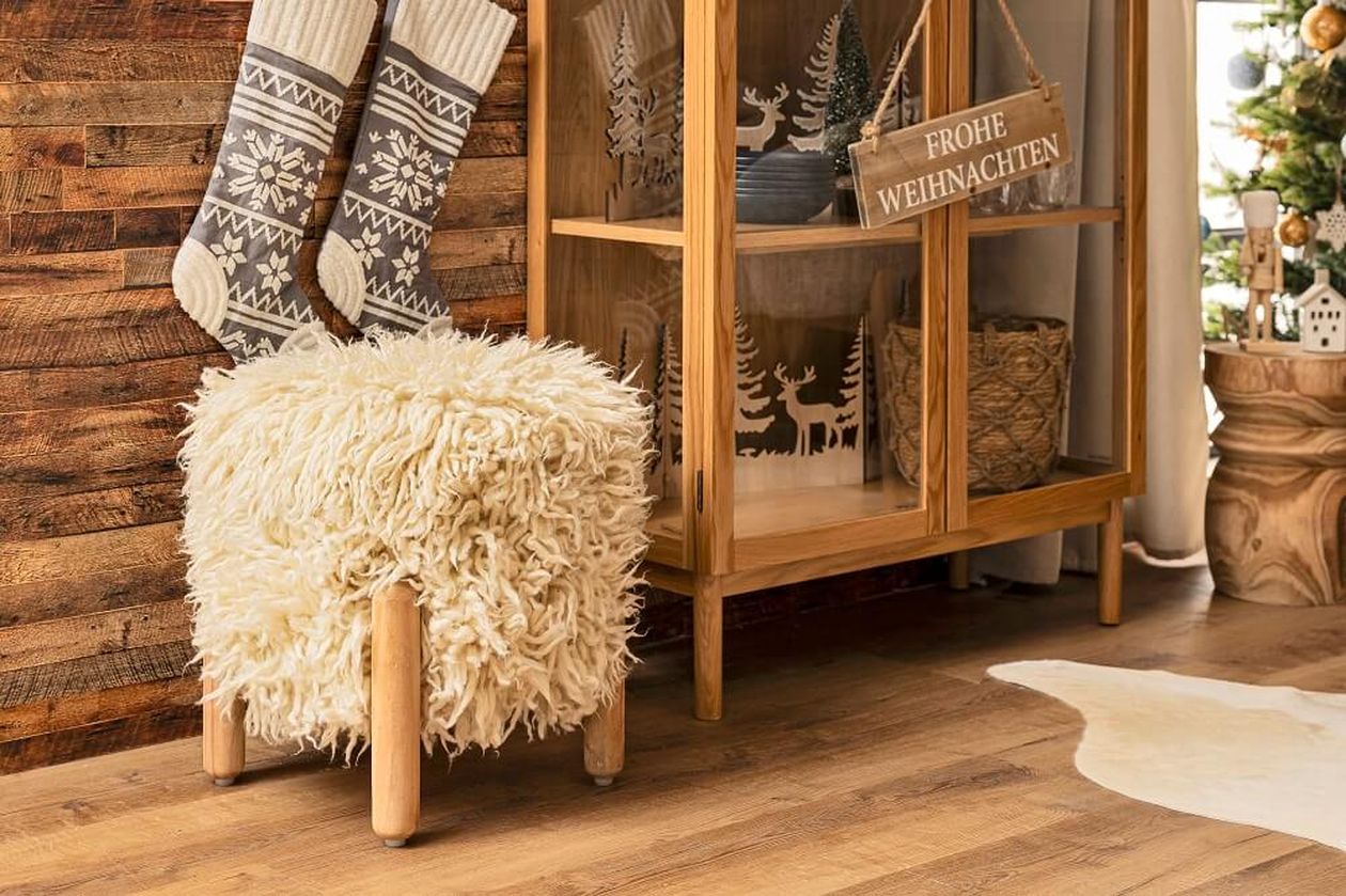 Gemütliches Zimmer mit flauschigem Hocker, Holzschrank, Strümpfen und „Frohe Weihnachten“-Schild. Weihnachtsbaum rechts teilweise sichtbar.