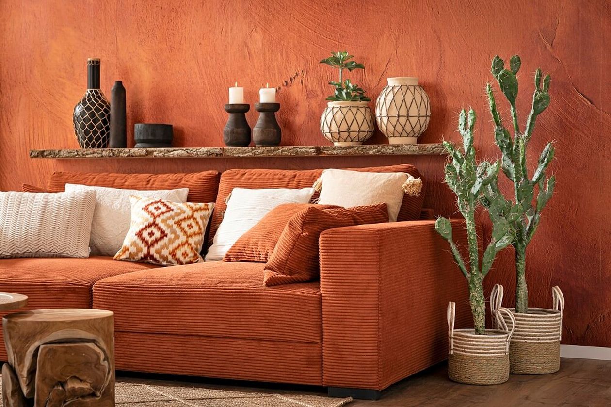 Gemütliches Wohnzimmer mit einem rostfarbenen Sofa, verschiedenen Kissen, einem Topfkaktus, geflochtenen Töpfen und Dekorationsgegenständen vor einer strukturierten orangefarbenen Wand.