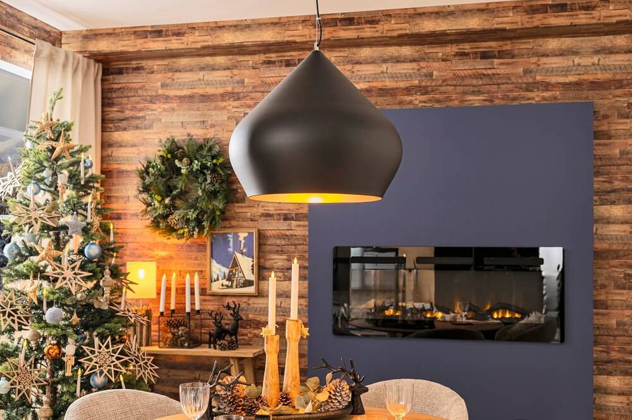 Gemütliches Wohnzimmer mit einer modernen Hängelampe, einem Weihnachtsbaum, einem Kranz, Kerzen und einem brennenden Kamin an einer Akzentwand aus Holz.