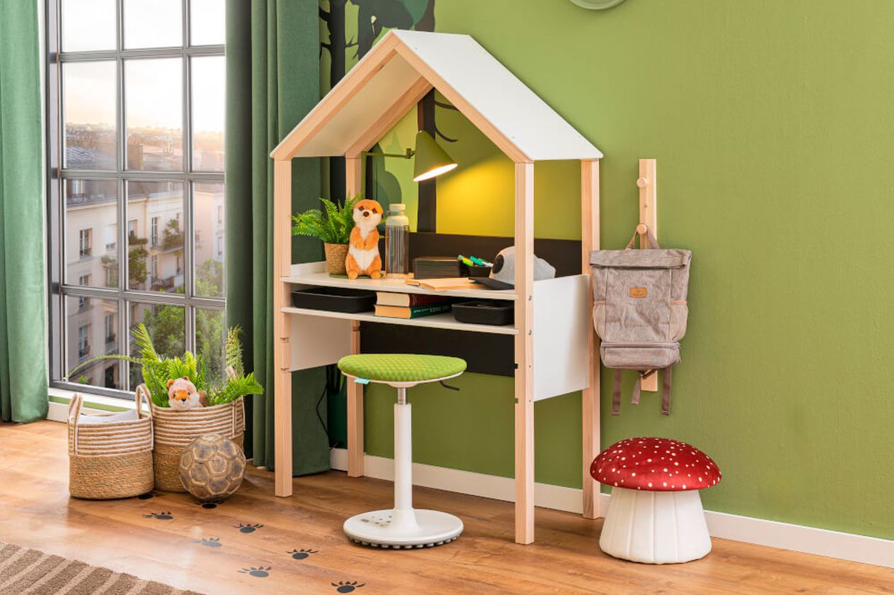 Kinderschreibtisch mit hausförmiger Platte, grünem Hocker, Plüschtieren und einem Pilzhocker in einem Zimmer mit grünen Wänden und großem Fenster.