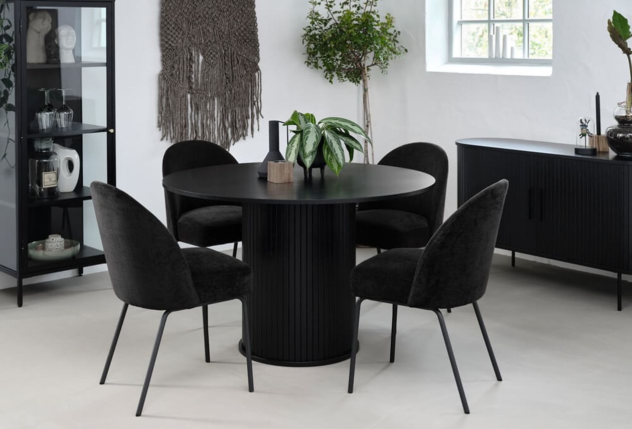 Runder schwarzer Esstisch mit geriffeltem Säulenfuß, umgeben von vier schwarzen Polsterstühlen mit Metallbeinen. Der Tisch steht in einem hellen Esszimmer mit weißen Wänden, einer Vitrine, einem Sideboard und Dekoelementen wie Pflanzen und Makramee-Wandbehang.