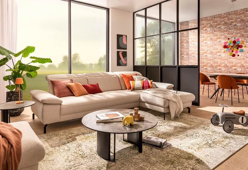 Helles Ecksofa in einem modernen Wohnzimmer mit großen Fenstern, Beistelltischen, bunten Kissen und einer Glas-Raumtrennung zu einem Essbereich mit Backsteinwand.