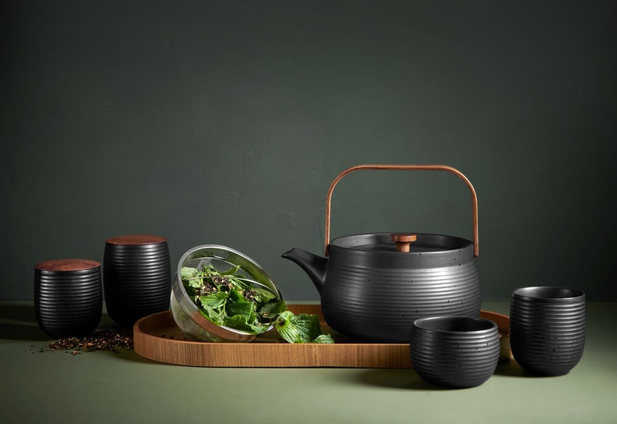 Schwarzes Teeservice im Japandi-Stil mit gerillter Kanne mit Holzgriff, Teebecher und Vorratsdosen auf einem Holztablett, dazu frische Kräuter.