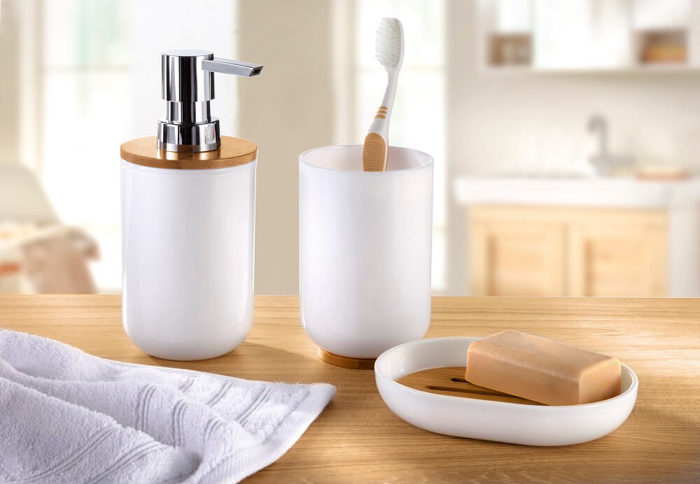 Badezimmerzubehörset mit einem Seifenspender, einem Zahnbürstenhalter und einer Seifenschale auf einer Holzablage, mit einem Handtuch in der Nähe.