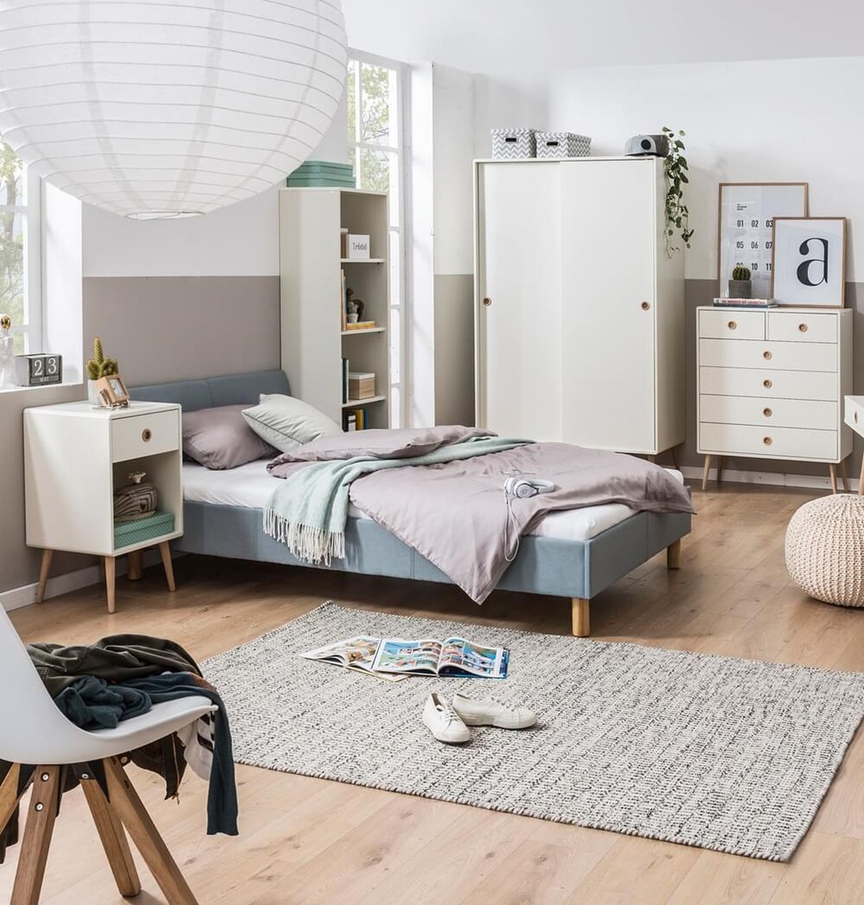 Graues Polsterbett mit gepolstertem Kopfteil in einem skandinavisch eingerichteten Jugendzimmer mit weißen Möbeln, Schrank und Schreibtisch.