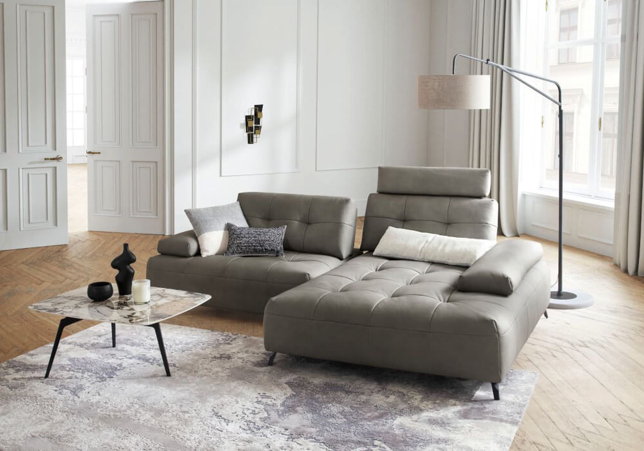 Ein modernes Wohnzimmer mit einem grauen Ecksofa, einem Couchtisch aus Marmor und einer Stehlampe. Durch die großen Fenster fällt Licht herein und bringt den Parkettboden zur Geltung.