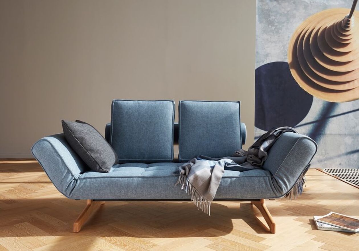 Flexibles Schlafsofa mit blauem Bezug und seitlich hochklappbaren Armlehnen sowie Holzbasis im Sonnenlicht.