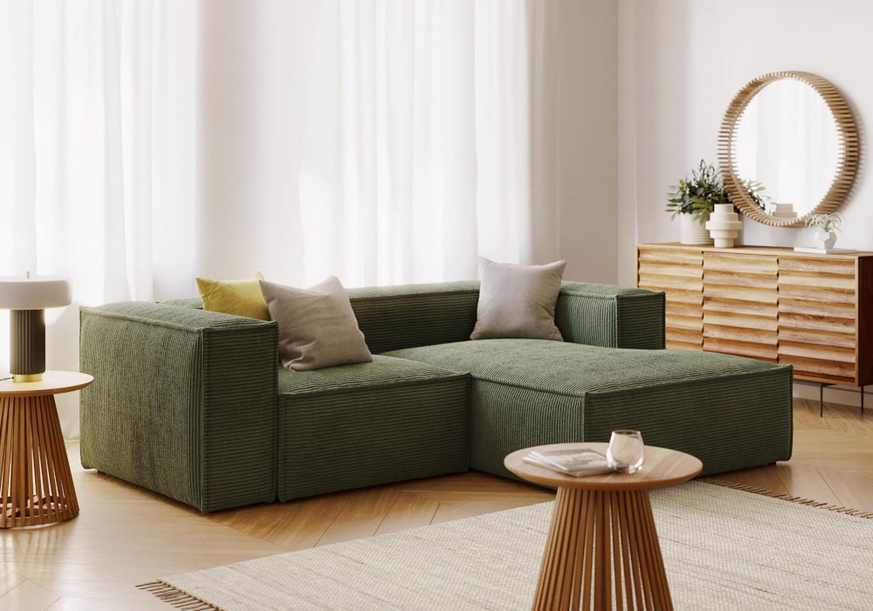 Ein gemütliches Wohnzimmer mit einem grünen Ecksofa, Beistelltischen aus Holz, einem runden Spiegel und einer Kommode aus Holz, das von sanftem Tageslicht erhellt wird.