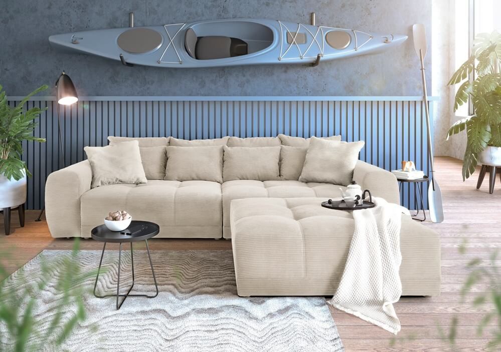 Großes cremefarbenes Cord-Sofa mit Hocker in einem maritim eingerichteten Wohnzimmer. An der Wand hängt ein graues Kajak, darunter eine blaue Holzlamellen-Verkleidung. Ein grau-weißer Wellenteppich und Beistelltische ergänzen das Ambiente.