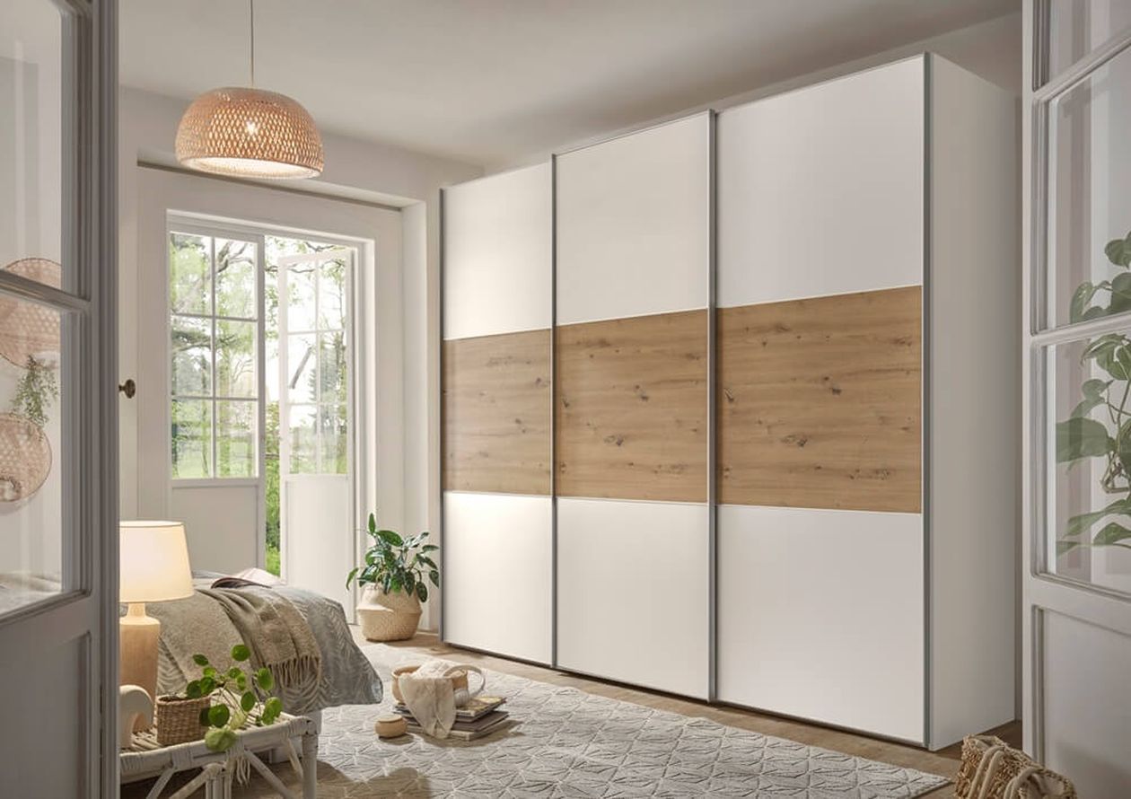 Gemütliches Schlafzimmer mit einem großen Kleiderschrank aus Holz und weißen Paneelen, einem Bett mit einer gestrickten Decke, Pflanzen und einem Fenster, das natürliches Licht hereinlässt.