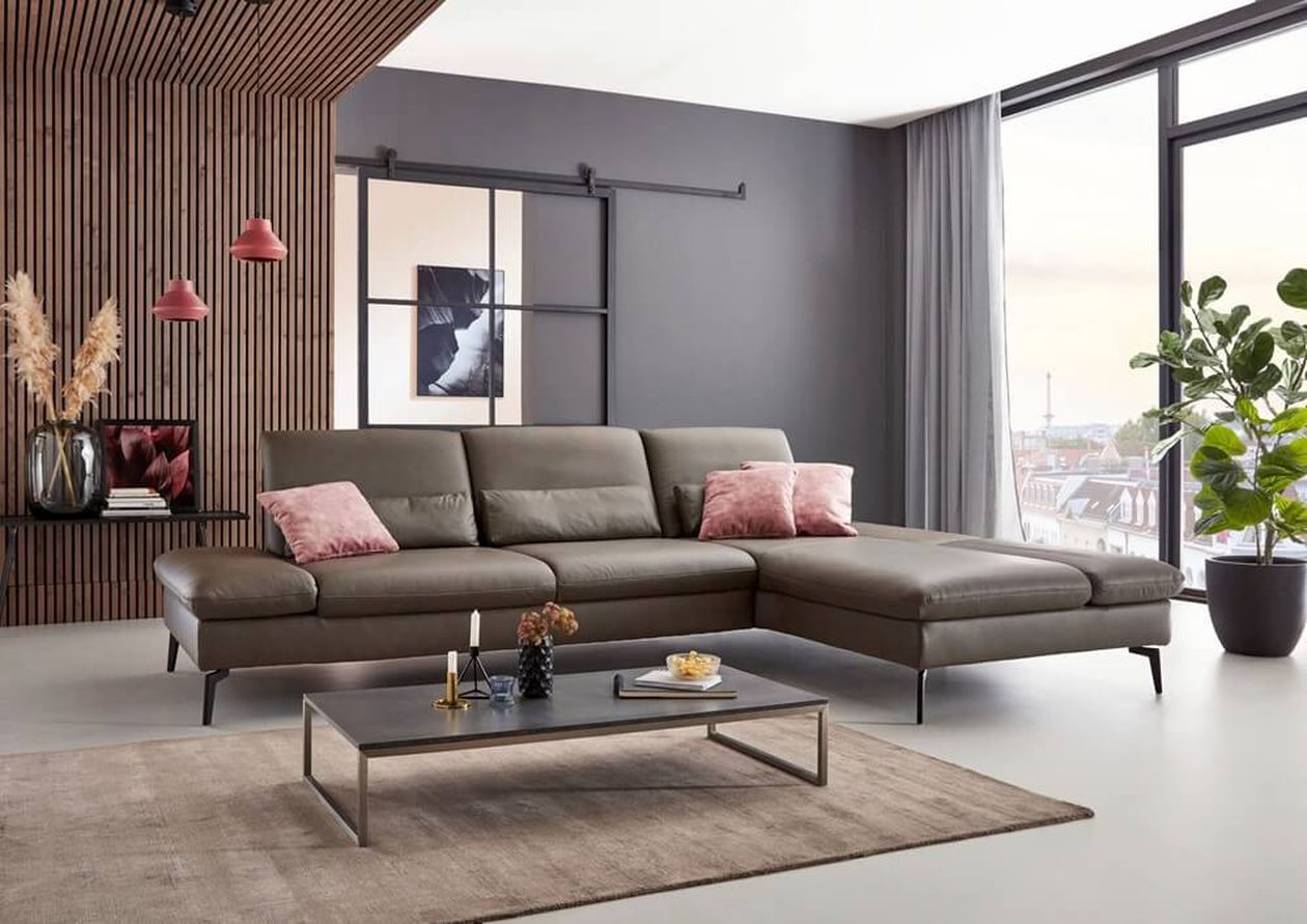 Modernes Wohnzimmer mit grauem Leder-Ecksofa mit Récamiere rechts und rosa Zierkissen. Dahinter eine Holzlamellenwand, eine Schiebetür und roséfarbene Hängelampen. Vor dem Sofa ein flacher Couchtisch aus Metall. Große Panoramafenster mit Blick auf die Stadt.
