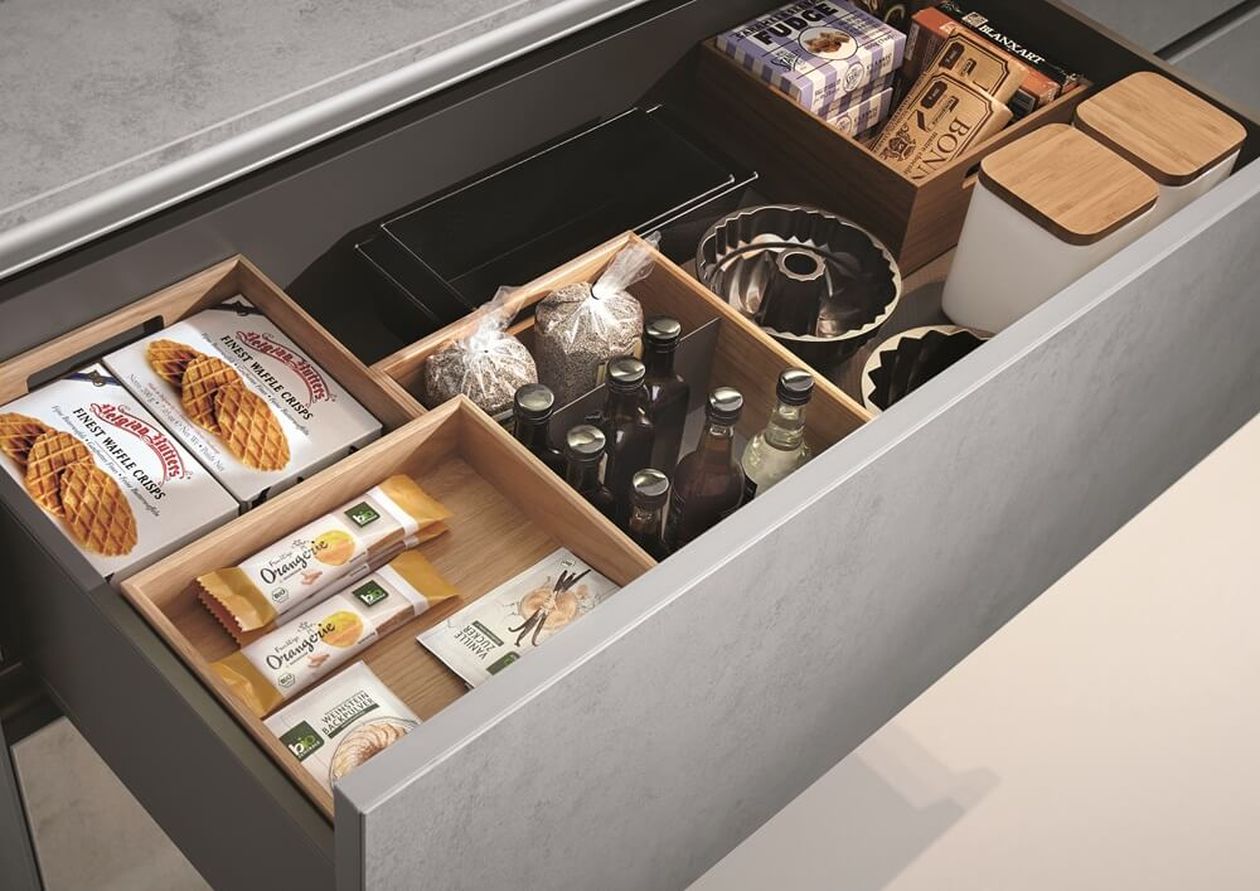 Offene Küchenschublade mit Snacks, Getränken und Backwaren, darunter abgepackte Waffeln, Kekse, Getränkeflaschen und Backformen.