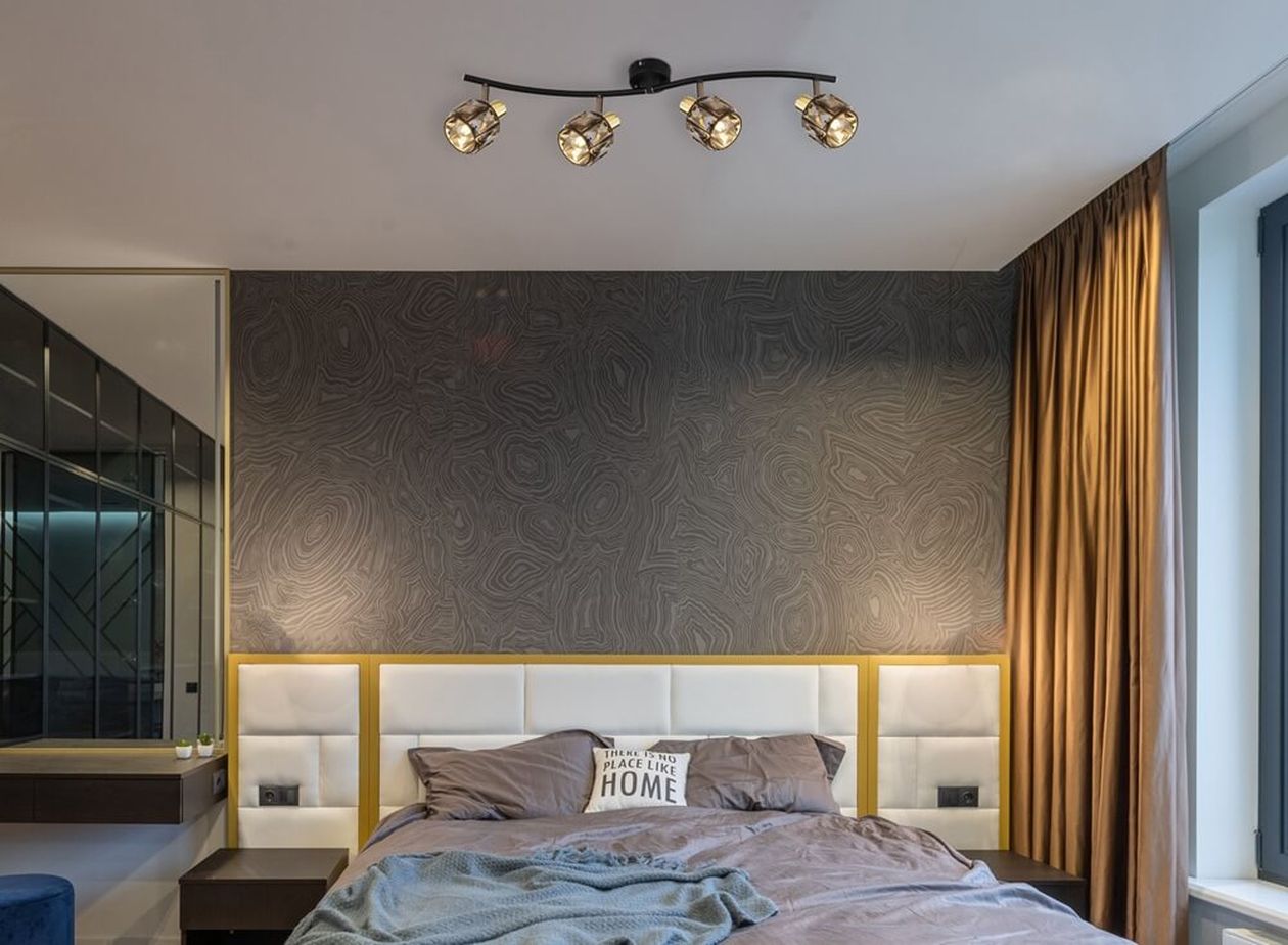 Vier Glasstrahler sind an einer geschwungenen Deckenleiste über einem Bett mit heller Kopflehne montiert.