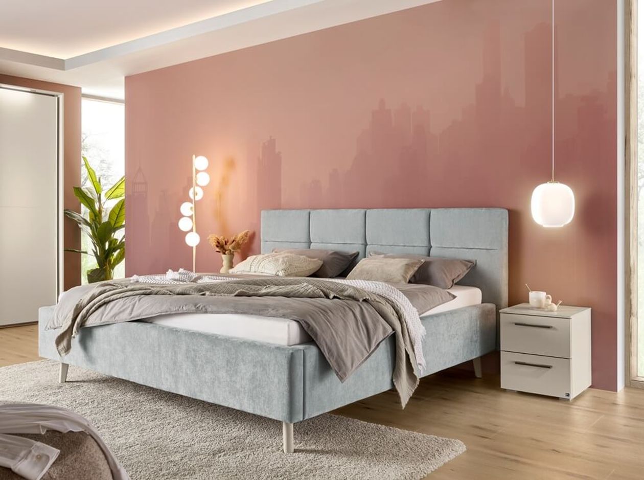 Graues Polsterbett steht vor einer rosafarbenen Wand mit einer modernen Stehleuchte und einer Hängelampe.
