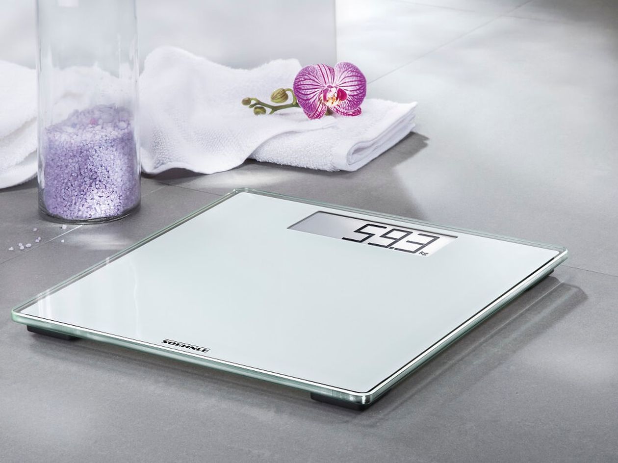 Eine digitale Personenwaage zeigt 59,3 kg auf einem grauen Fliesenboden an; im Hintergrund befinden sich eine lila Orchidee und ein Handtuch.