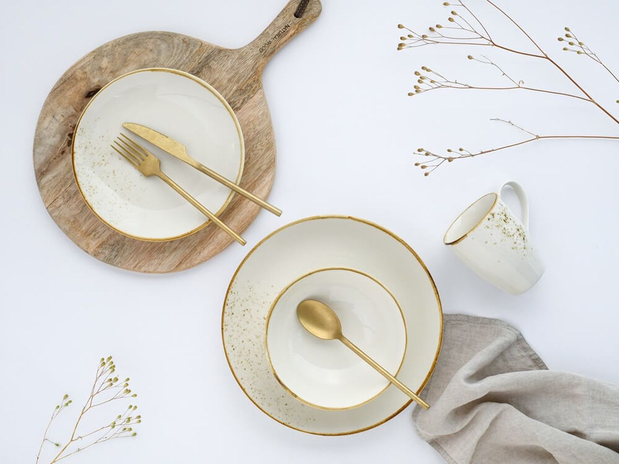 Weißes Porzellan-Kombiservice mit cremefarbenen Tellern und goldenen Sprenkeln sowie Goldrand, mit mattem Goldbesteck auf Holzbrett und weißem Untergrund, dekoriert mit Trockenblumenzweigen und einer Leinenseviette.