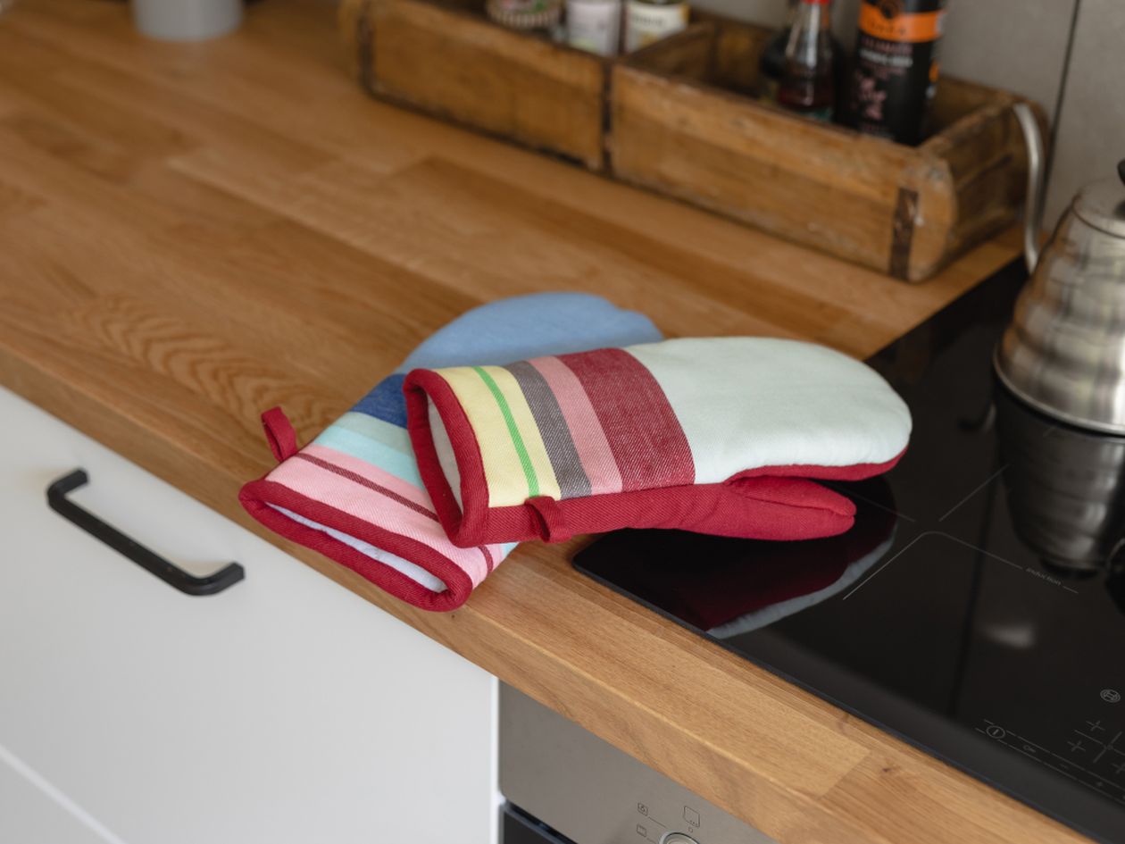 Die beiden Handschuhe des Ofenhandschuh Sets Nr. 1 von Remember Liegen auf eine hölzernen Arbeitsplatte. Sie haben ein schickes Design aus bunten Streifen.