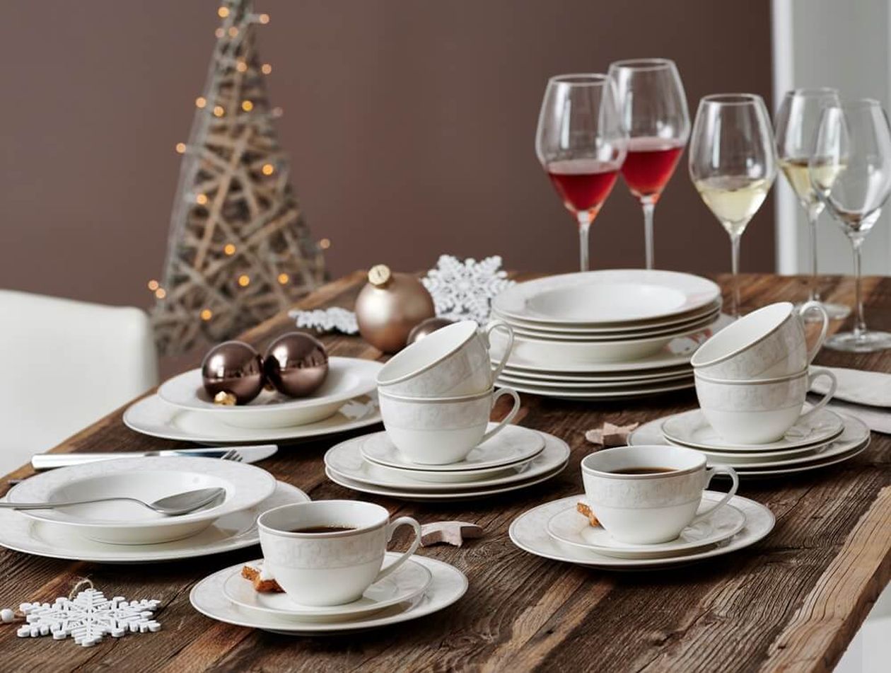 Elegantes Tischgedeck mit weißem Porzellan, Kaffeetassen, Weingläsern und Weihnachtsdekor, darunter ein Holzbaum und Ornamente, auf einem Holztisch.