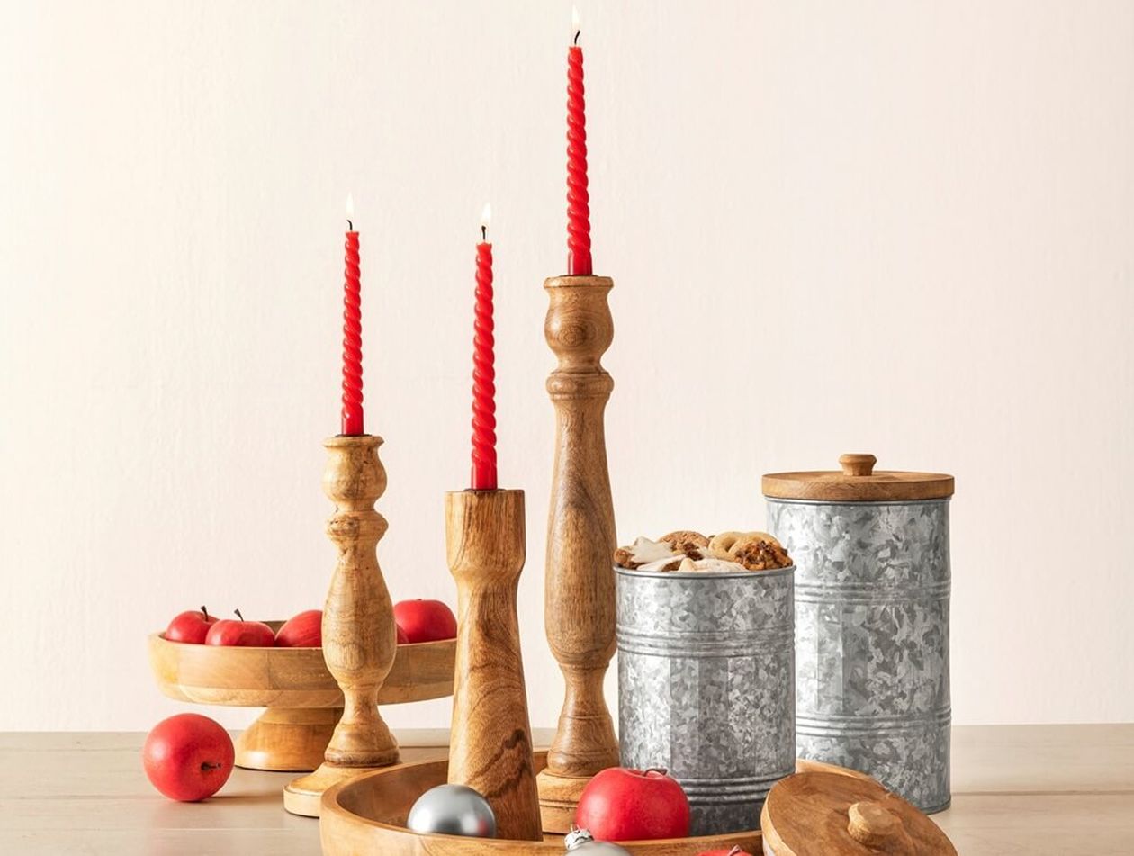 Kerzenständer aus Holz mit roten Kerzen, Metallbehälter und Schalen mit Äpfeln und Ornamenten auf einer hellen Oberfläche.