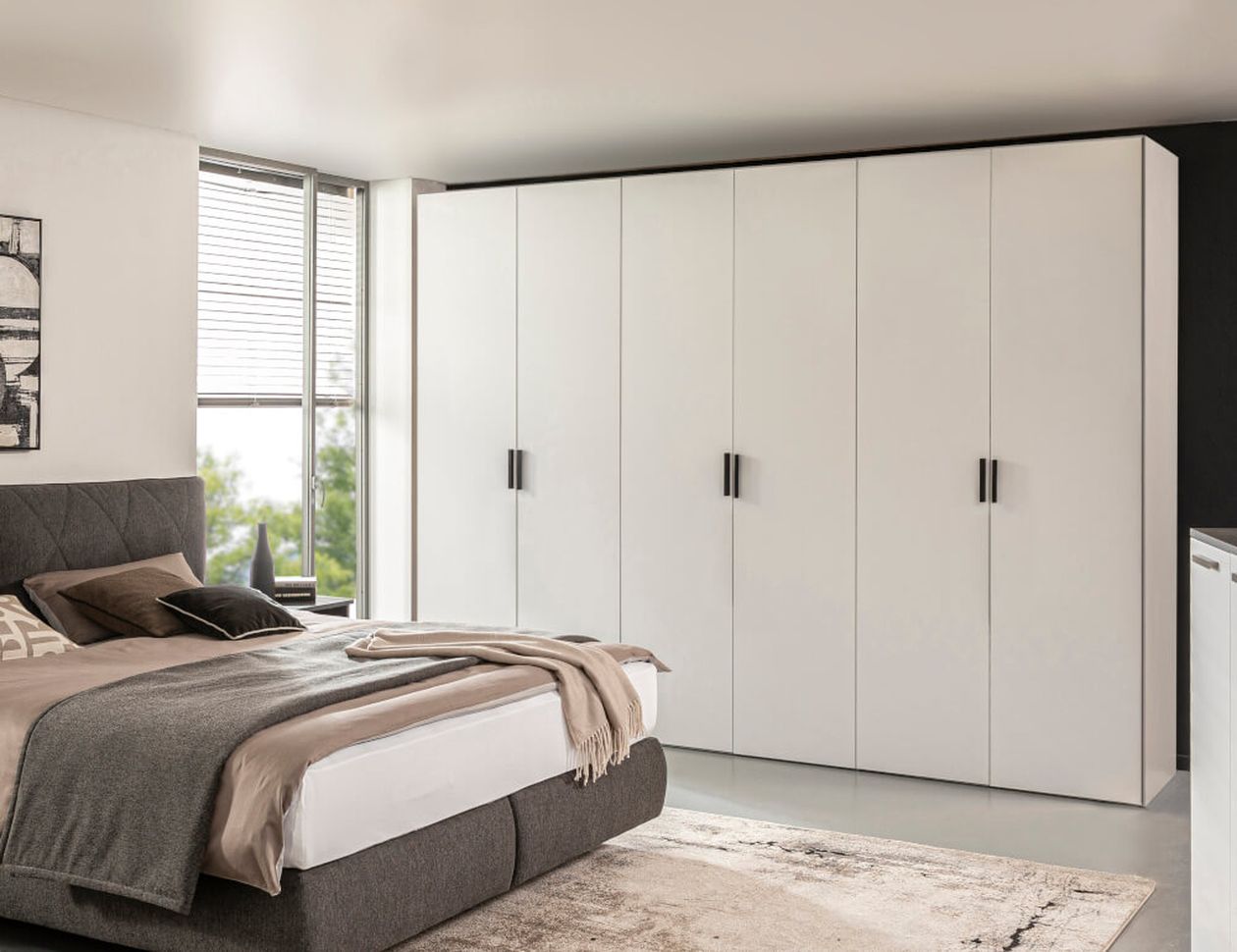 Modernes Schlafzimmer mit großem weißen Kleiderschrank, grauem Polsterbett und weicher Bettwäsche. Sonnenlicht strömt durch ein Fenster auf der linken Seite.