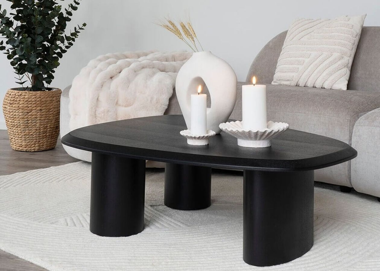 Schwarzer ovaler Couchtisch mit drei zylindrischen Beinen, dekoriert mit weißen Kerzen und einer organisch geformten Vase, vor einem grauen Sofa mit strukturierten Kissen.