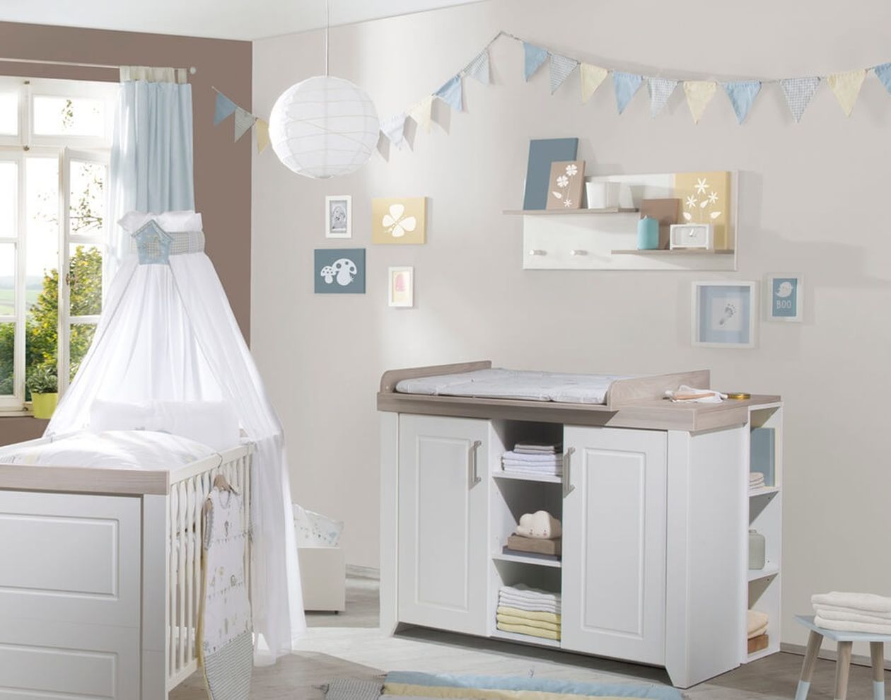 Helles Babyzimmer mit weißer Wickelkommode mit zwei Türen, Wickelaufsatz und seitlichem Regal, daneben ein weißes Babybett mit weißem Baldachin und Wimpelkette an der Wand.