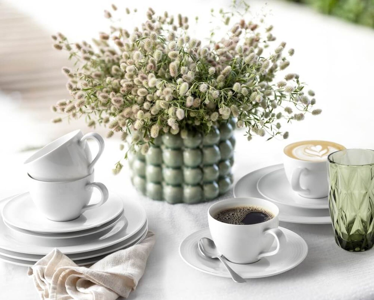 Weißes Kaffeeservice mit gestapelten Tassen und Tellern sowie zwei gefüllten Kaffeetassen auf weißem Tischtuch, daneben ein grünes Glas und eine grüne Bubble-Vase mit flauschigen Trockenblumen.