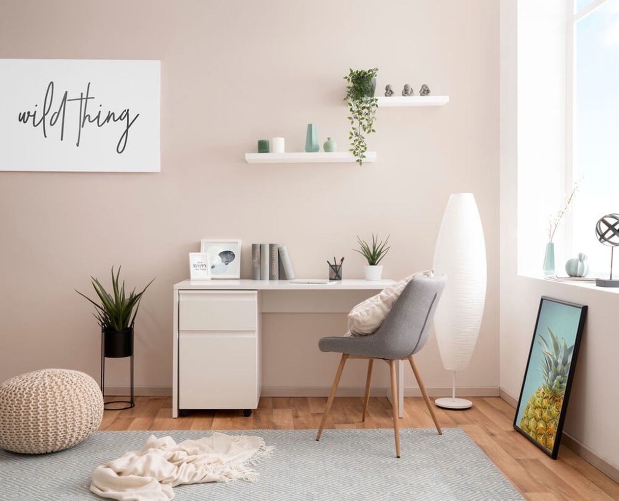 Minimalistisches Homeoffice mit weißem Schreibtisch, grauem Stuhl, Pflanzen und Deko. Wandbild mit dem Schriftzug „Wild Thing“. Sanfte Beleuchtung durch eine hohe Lampe.