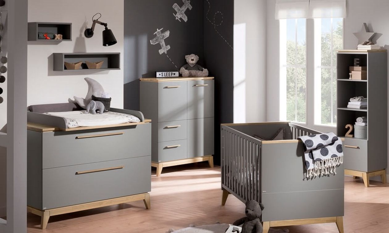 Modernes Babyzimmer in Grau mit Holzakzenten, bestehend aus Wickelkommode, Babybett mit Sprossen, Highboard und Regal. An der dunkelgrauen Wand Flugzeug-Wanddekoration. Graue Wandregale und eine schwarze Wandleuchte ergänzen die Einrichtung.