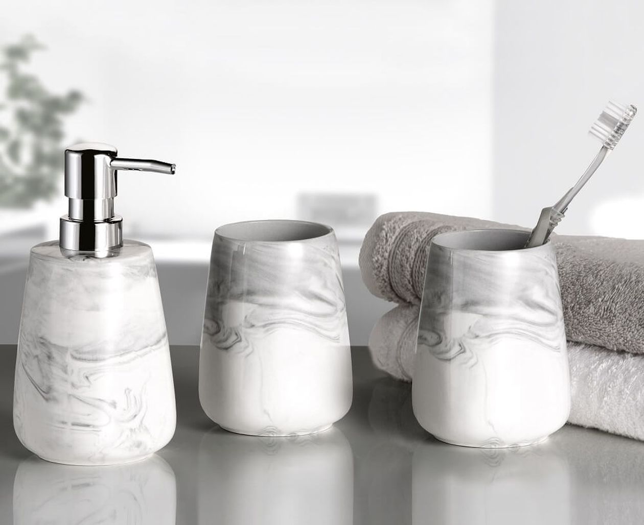 Seifenspender mit Marmor-Muster, zwei Becher und eine Zahnbürste neben aufgestapelten Handtüchern auf einer reflektierenden Oberfläche.