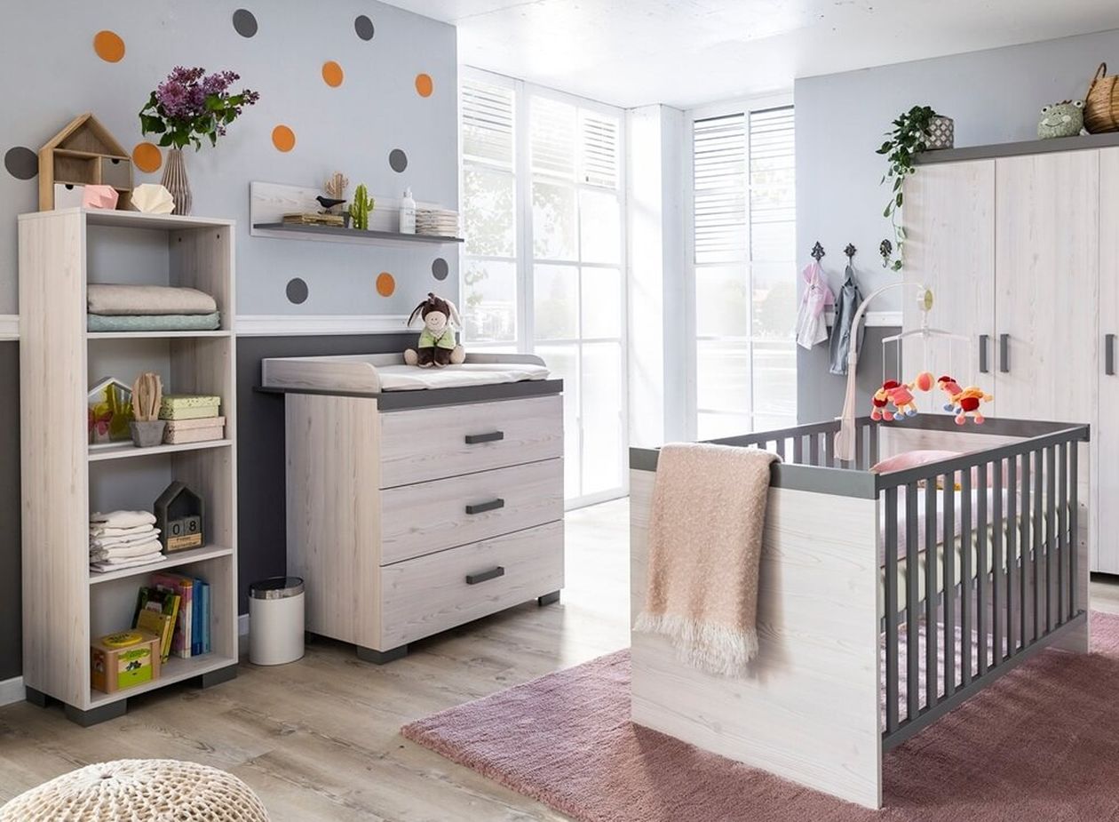 Helles Babyzimmer mit weiß-grauem Möbelset aus Wickelkommode, Schrank, Regal und Babybett; bunte Wandpunkte in Orange und Grau an der grauen Wand, rosa Teppich auf dem Boden.
