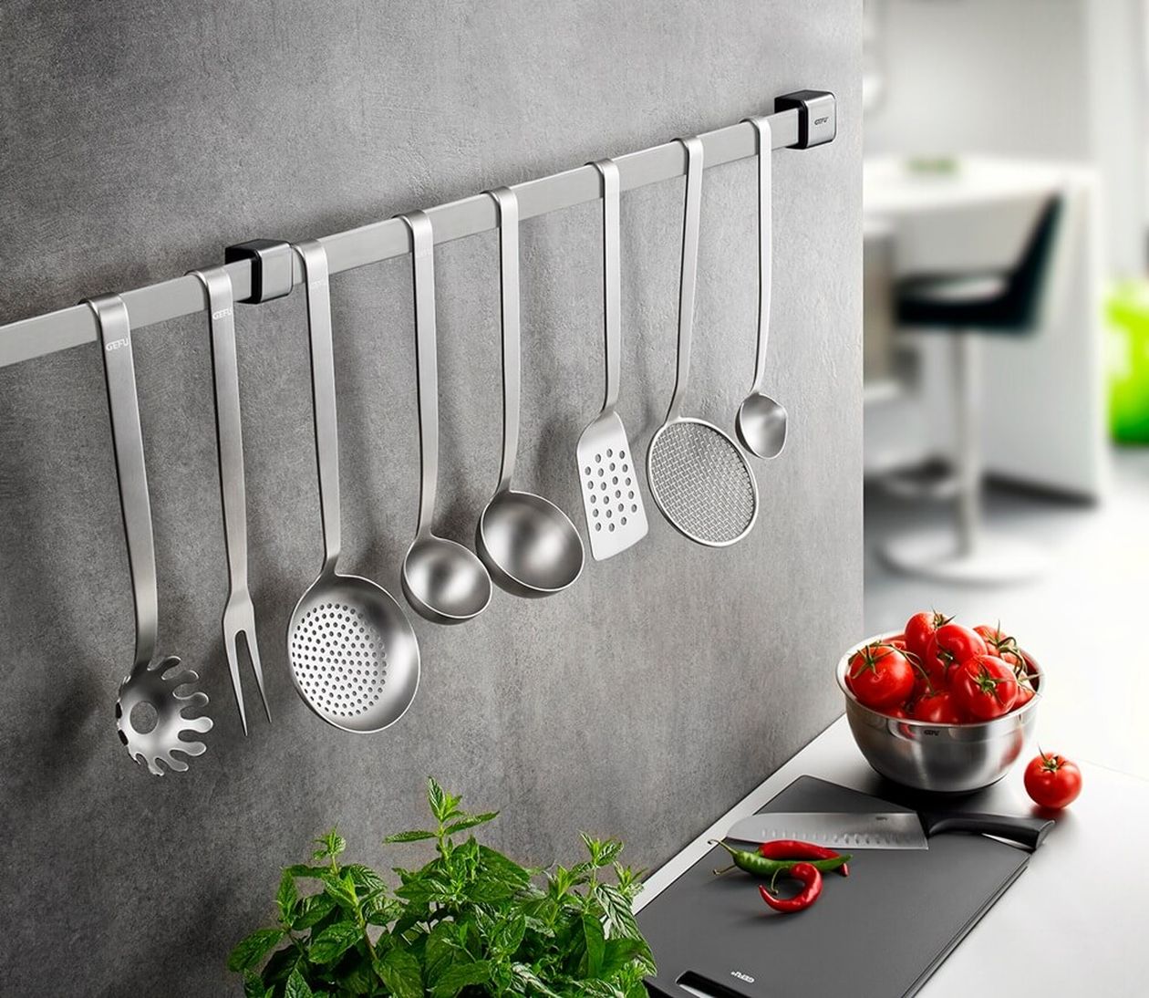 An einer Edelstahlleiste an der Küchenwand hängen verschiedene Küchenhelfer aus Metall. Darunter stehen frische Kräuter, Tomaten und Chilis auf einer Arbeitsfläche.