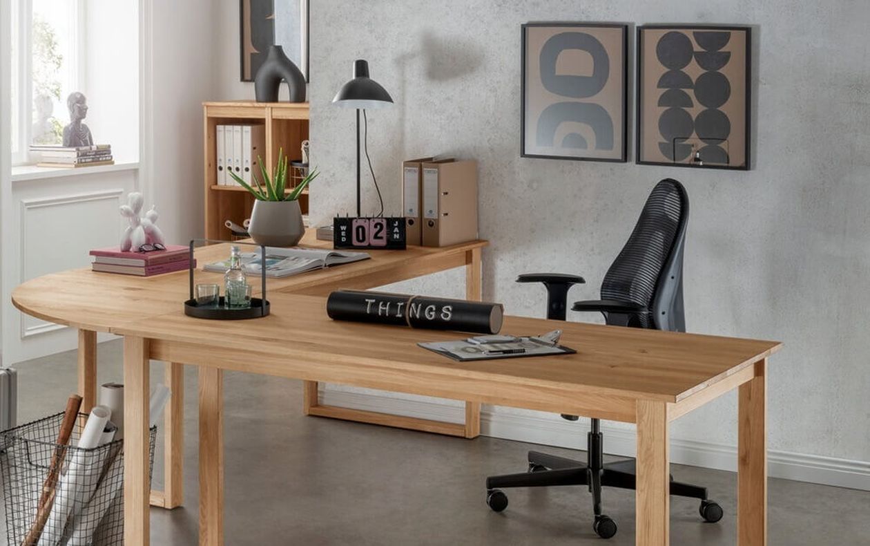 L-förmiger Schreibtisch aus massivem Eichenholz mit Kufengestell in einem modernen Home-Office mit grauer Betonwand, schwarzem Bürostuhl und Holzregal.