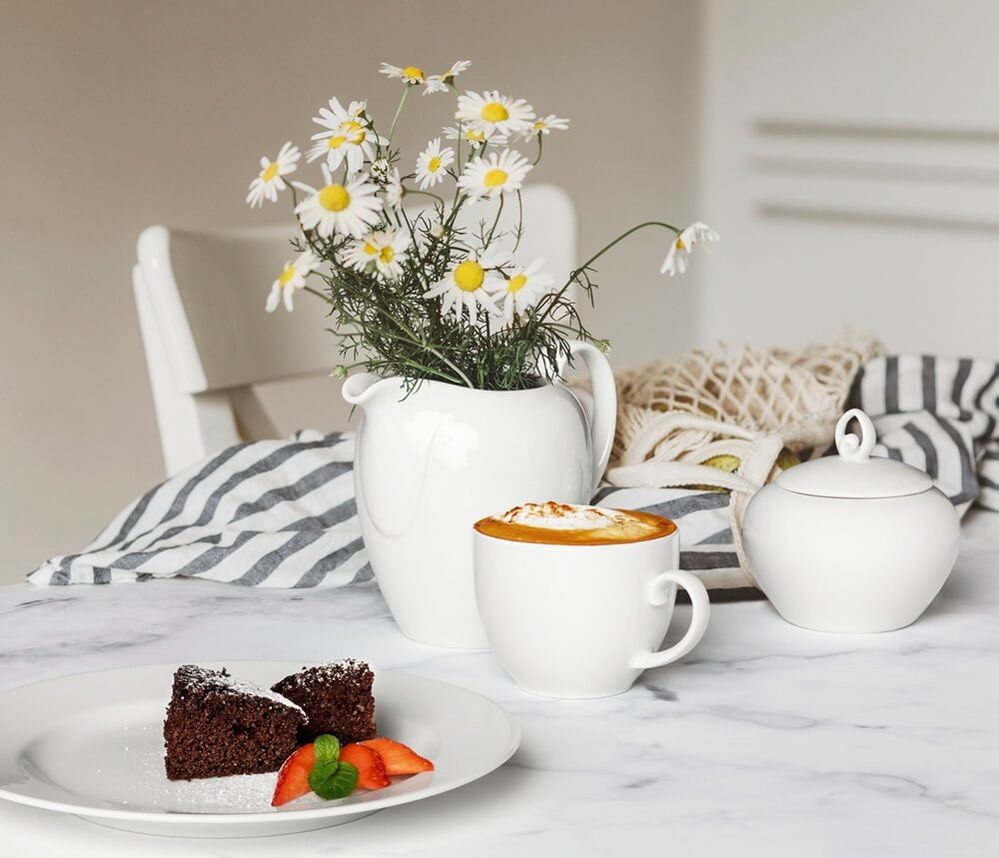 Weißer Frühstücksteller mit geschwungenem Rillenrand, einer Kaffeetasse und Zuckerdose auf einem Marmortisch, daneben ein Cappuccino, ein Stück Schokoladenkuchen mit Erdbeeren und ein Blumenstrauß aus Margeriten in einer weißen Kanne.