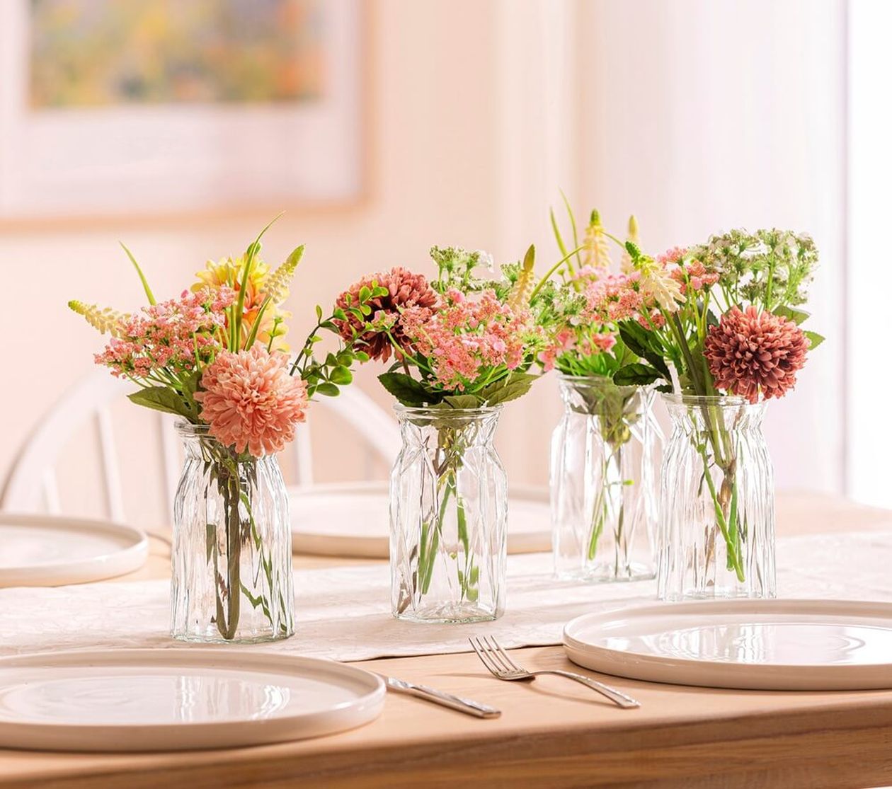 Vier Glasgefäße mit rosa und grünen Blumen auf einem Esstisch mit weißen Tellern und Besteck in einem sanft beleuchteten Raum.