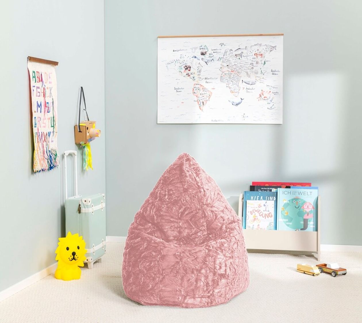 Kinderzimmer mit einem rosa Sitzsack, einer Weltkarte an der Wand, Büchern in einem Regal, einem kleinen Koffer und einer gelben, löwenförmigen Lampe.