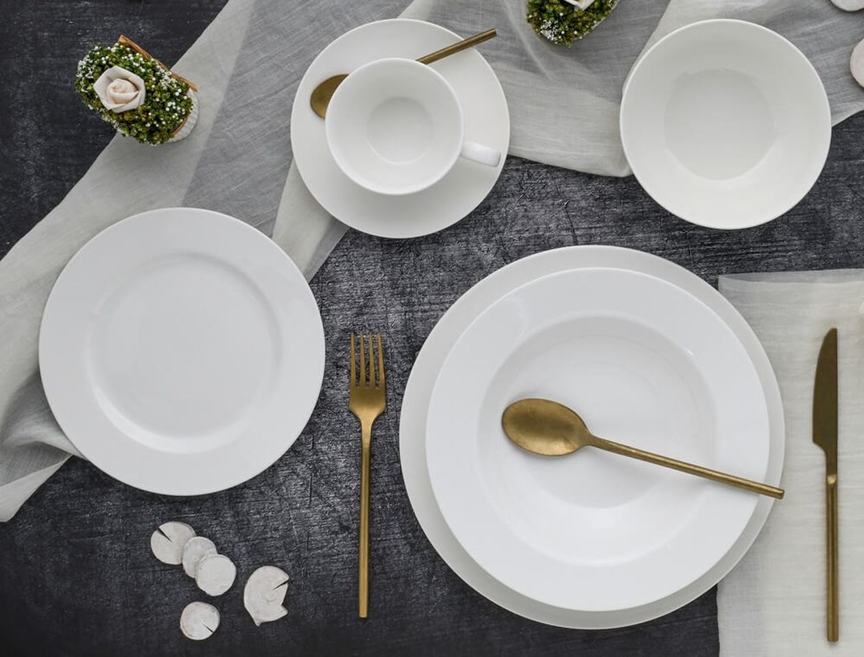 Gedeckter Tisch von oben mit weißem Kombiservice aus Teller, Schalen und Tasse, mattgoldenem Besteck, weißen Rosendekorationen und grauer Leinenserviette auf dunklem Untergrund.