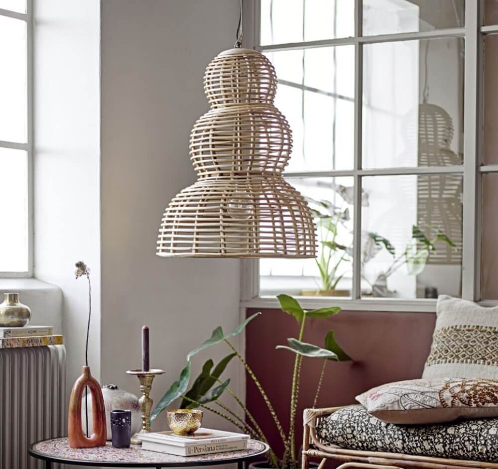 Ein gemütliches Zimmer mit einer Pendelleuchte aus Korbgeflecht, einem kleinen Tisch mit Dekor, Pflanzen und einem Sofa mit gemusterten Kissen in der Nähe großer Fenster.
