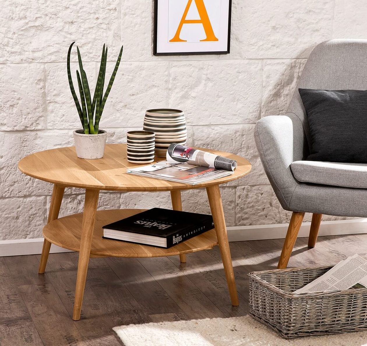 Ein gemütliches Wohnzimmer mit einem runden Couchtisch aus Holz, einer Topfpflanze, gestreiften Tassen, Zeitschriften und einem grauen Sessel auf einem Holzboden.