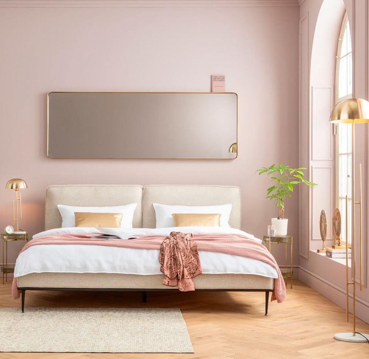 Elegantes Schlafzimmer mit beigem Bett, rosa Überwurf, großem Spiegel, goldenen Lampen und einer Topfpflanze am Bogenfenster. Sanfte, warme Beleuchtung.