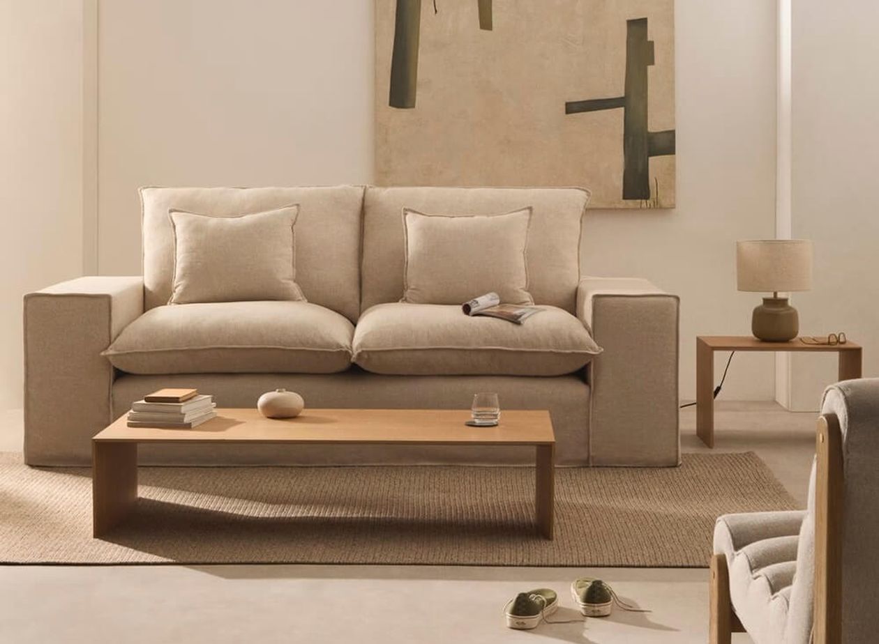 Beiges Leinensofa mit tiefen Kissen und breiten Armlehnen in hellem Wohnzimmer mit abstraktem Wandbild und schlichtem Holz-Couchtisch.