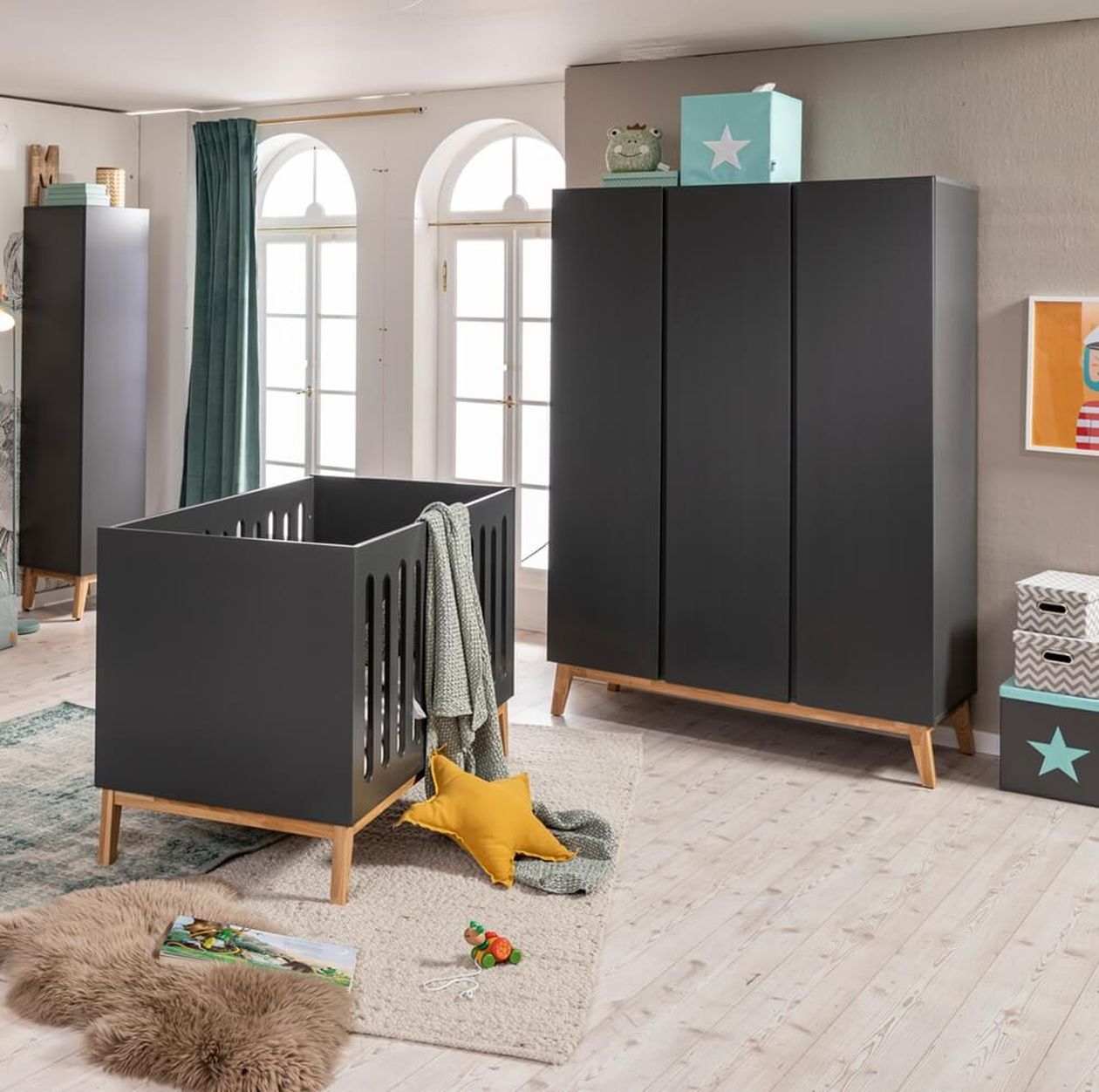 Dunkelgrau-schwarzes Babybett mit Holzfüßen und Bogenausschnitten an den Seiten, daneben ein passender dreitüriger Kleiderschrank; gelbes Sternkissen und grüne Decke als Deko, heller Holzboden und mintgrüne Akzente im Hintergrund.