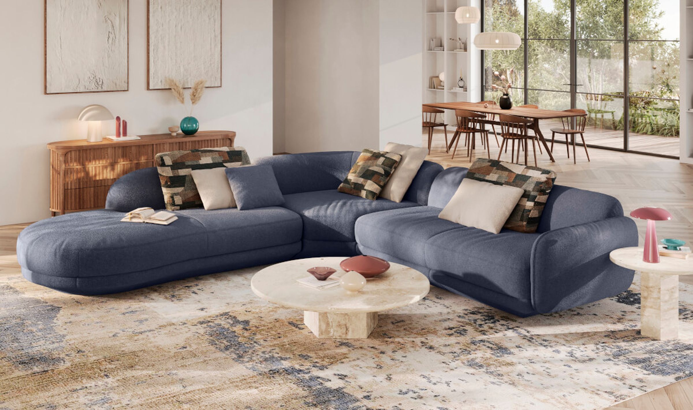 Ein modernes Wohnzimmer mit einem blauen Sofa, gemusterten Kissen, einem runden Couchtisch aus Marmor und einem Essbereich im Hintergrund.