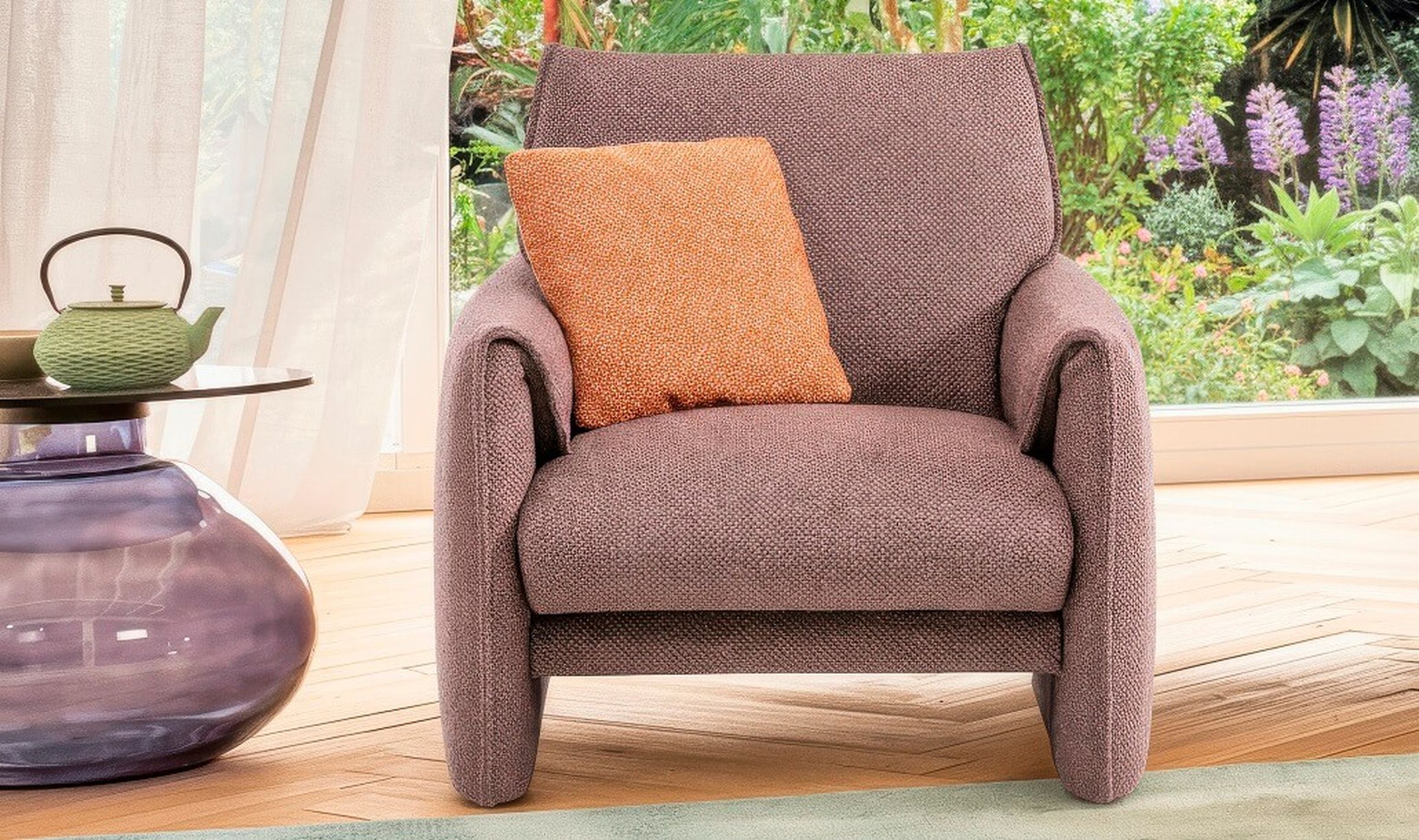 Ein gemütlicher brauner Sessel mit einem orangefarbenen Kissen auf einem Holzboden, daneben ein Glastisch und eine Teekanne und im Hintergrund ein Blick auf den Garten.