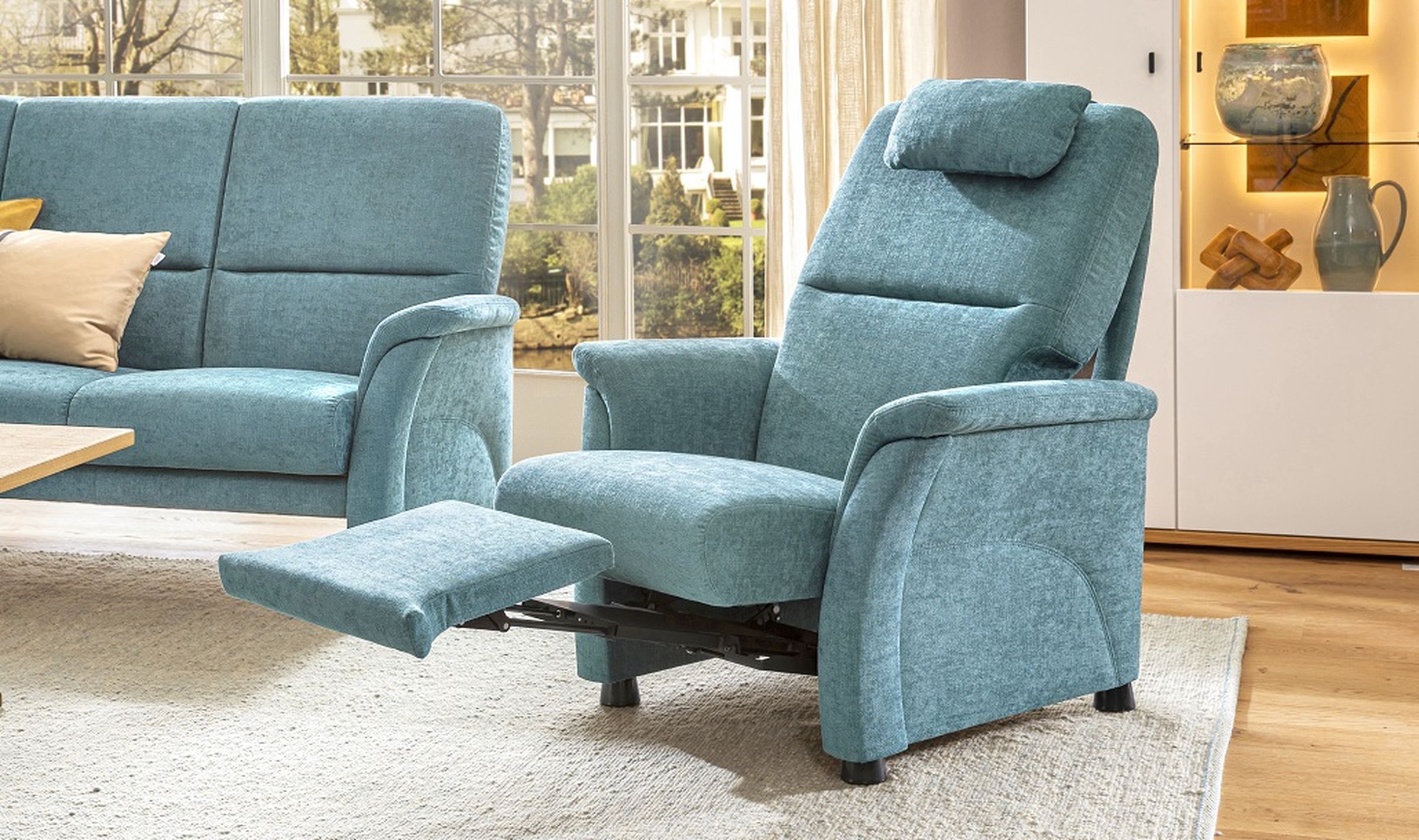 Ein blaugrüner Relaxsessel mit verlängerter Fußstütze in einem sonnendurchfluteten Wohnzimmer, neben einem passenden Sofa und einem Holzschrank mit Dekoartikeln.